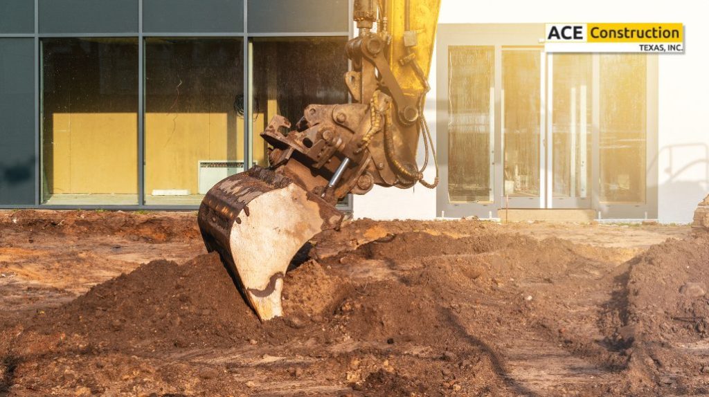A construction site featuring an excavator's bucket digging into the soil, with a glass storefront in the background.