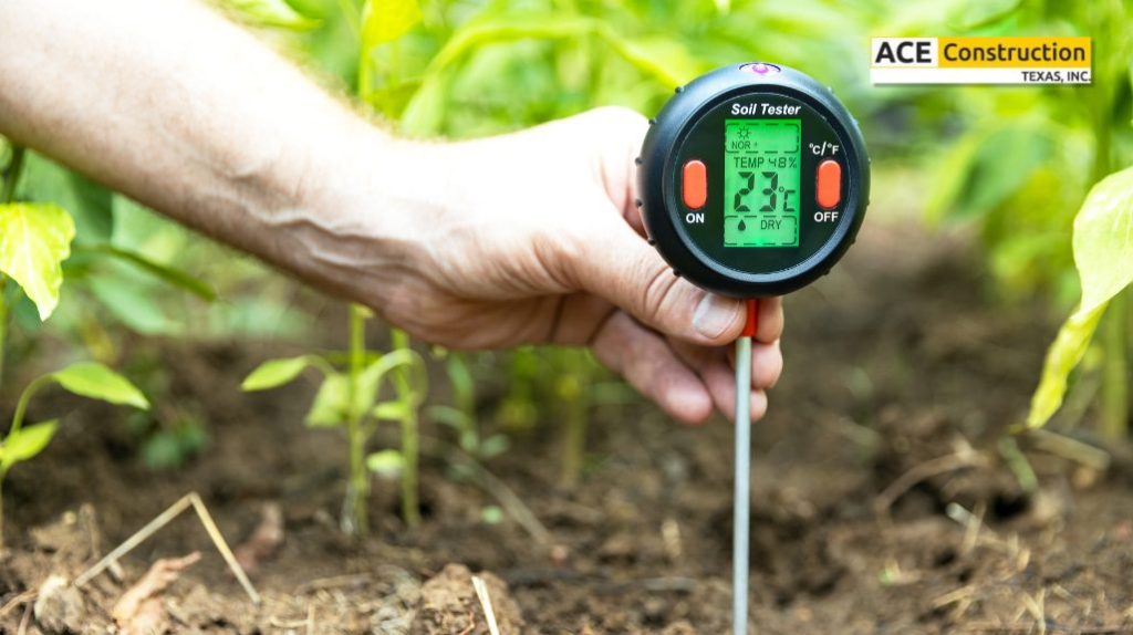 A hand holds a digital soil tester in a garden, displaying temperature and moisture levels among green plants.